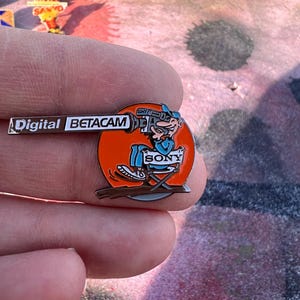 Pode incluir: Um pin vintage da Sony Digital Betacam. O pin apresenta um personagem de desenho animado operando uma câmera, em um círculo laranja. O pin tem o logotipo da Sony e as palavras "Digital Betacam".
