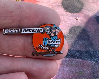 May include: A vintage Sony Digital Betacam pin. The pin features a cartoon character operating a camera, set against an orange circle. The pin has the Sony logo and the words "Digital Betacam".