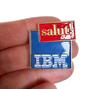Könnte beinhalten: Eine rechteckige Emaille-Anstecknadel mit einer blauen quadratischen Basis und dem weiß-blau gestreiften IBM-Logo. Ein kleinerer roter rechteckiger Bereich befindet sich oben rechts und enthält den goldfarbenen Text salut! mit Ausrufezeichen.