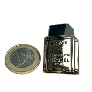 Puede incluir: Pin de metal dorado con forma de frasco de perfume. El frasco es negro y tiene el texto "EGOISTE" y "CHANEL PARIS" en &eacute;l. El texto "EAU DE TOILETTE" tambi&eacute;n est&aacute; en el frasco.