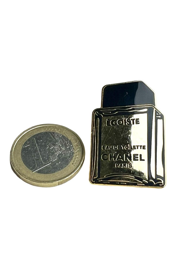 Puede incluir: Pin de metal dorado con forma de frasco de perfume. El frasco es negro y tiene el texto "EGOISTE" y "CHANEL PARIS" en &eacute;l. El texto "EAU DE TOILETTE" tambi&eacute;n est&aacute; en el frasco.