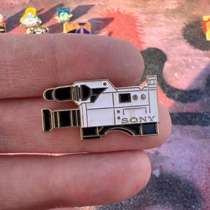Puede incluir: Un pin de esmalte blanco y negro con forma de cámara de video vintage, con la marca "SONY" impresa en el lateral. El pin tiene un borde dorado y está sobre un fondo colorido.