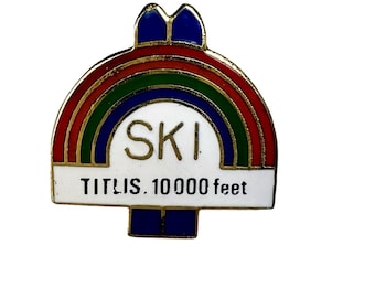 May include: A gold-toned metal enamel pin shaped like a semi-circle with a rainbow arch of red, green, and blue. The center features the word SKI in gold letters on a white background, with the text TITLIS. 10 000 feet printed in black below.