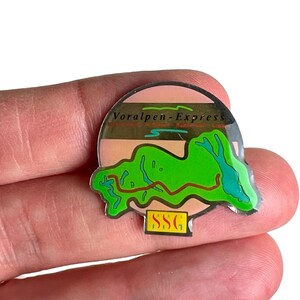 May include: A circular metal pin featuring a graphic of a green landscape with a brown line, a light pink sky background, and the text Voralpen-Express in black. At the bottom, a small yellow rectangular tab displays the black letters SSG.