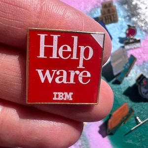 May include: A square, red pin with gold edging, displaying the white text "Helpware" and the IBM logo. The pin is held in a hand, with other pins visible in the background.
