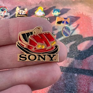Pode incluir: Um alfinete de esmalte vermelho e preto com um passeio de parque de diversões estilizado com a palavra "SONY" em letras douradas. O alfinete é um item de colecionador, provavelmente dos anos 1990 ou início dos anos 2000.