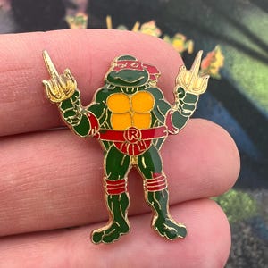May include: A green, red, and gold enamel pin of the Teenage Mutant Ninja Turtle Raphael, giving the middle finger with both hands. The pin has a gold outline and features the letter "R" on the belt.