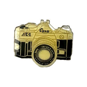 May include: Gold-colored metal pin of a Canon AE-1 camera with a black lens. The camera has a black background and the word "Canon" is printed in black on the top of the camera.
