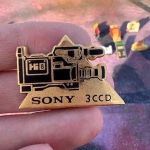 Puede incluir: Un pin dorado con un diseño de cámara de video vintage con detalles en negro. El pin tiene el texto "SONY 3CCD" y un logotipo "Hi 8". Este artículo de colección probablemente data de los años 80 o 90.