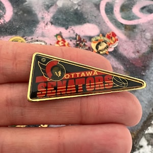 Puede incluir: Pin con forma de banderín de los Ottawa Senators con borde dorado, negro, rojo y blanco. El pin presenta el logotipo del equipo y las palabras "OTTAWA SENATORS". Un gráfico de disco de hockey está en el lado derecho.