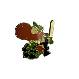 May include: A gold-toned metal enamel pin featuring a stylised cartoon character with a large brown head, a green hat, and a green patterned outfit. The character is holding a long, vertical gold-coloured sword-like object in a black hand.