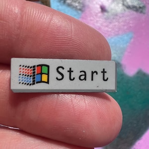 Puede incluir: Un pequeño botón rectangular con la palabra "Start" en texto negro. El botón presenta el clásico logotipo de Windows en la esquina superior izquierda, con un diseño colorido. El botón es gris y parece ser una tecla de ordenador.