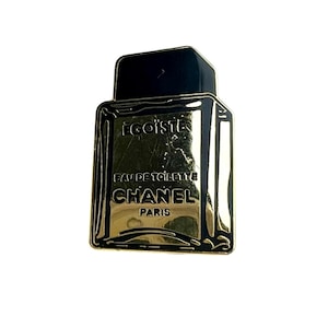 Puede incluir: Pin de metal dorado y negro con forma de frasco de perfume. El frasco dice "EGOISTE EAU DE TOILETTE CHANEL PARIS".