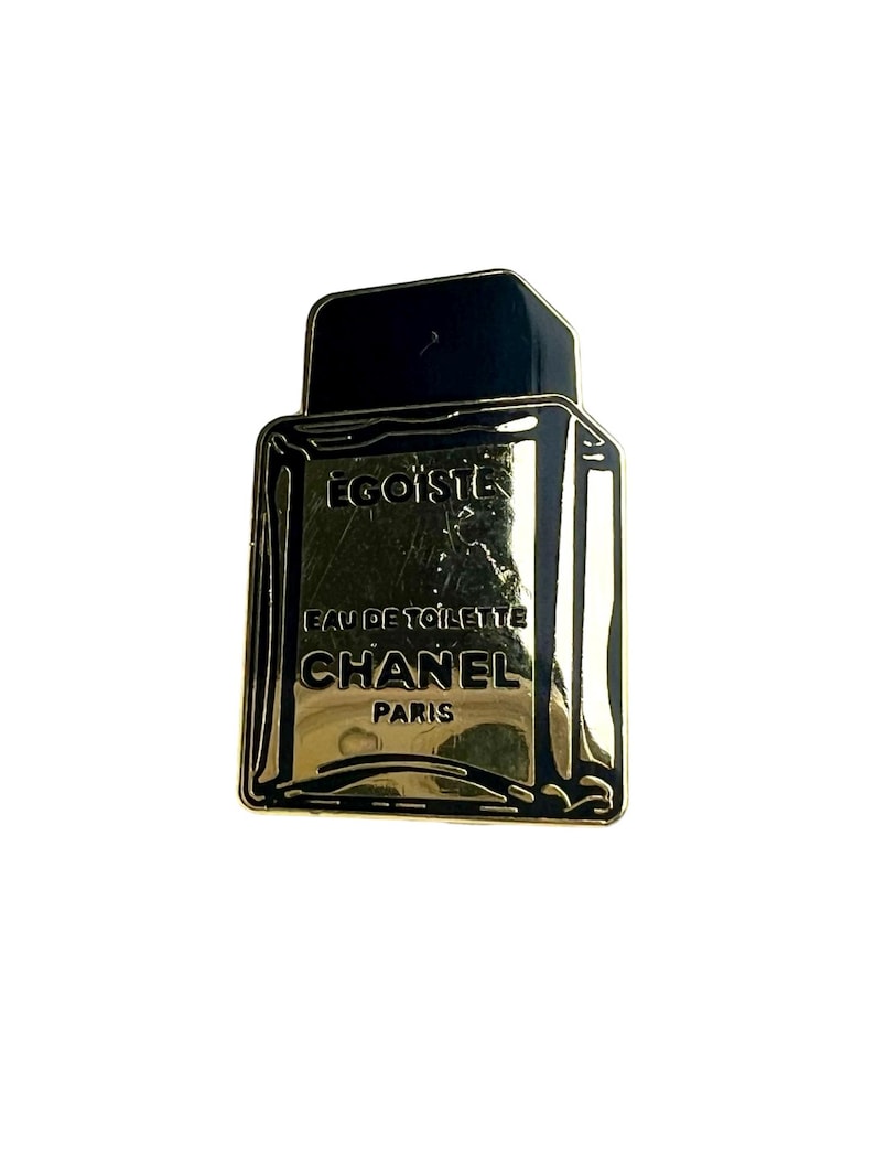 Puede incluir: Pin de metal dorado y negro con forma de frasco de perfume. El frasco dice "EGOISTE EAU DE TOILETTE CHANEL PARIS".
