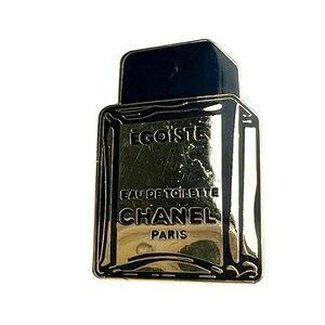 Puede incluir: Pin de metal de color dorado con forma de frasco de perfume. El frasco tiene una tapa negra y el texto "EGOISTE", "EAU DE TOILETTE", "CHANEL" y "PARIS" est&aacute; impreso en el frasco.