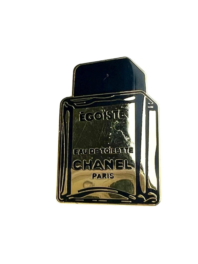 Puede incluir: Pin de metal de color dorado con forma de frasco de perfume. El frasco tiene una tapa negra y el texto "EGOISTE", "EAU DE TOILETTE", "CHANEL" y "PARIS" est&aacute; impreso en el frasco.