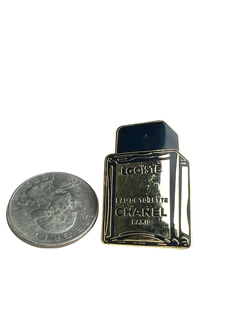 Puede incluir: Pin de metal dorado con forma de frasco de perfume. El frasco es negro y tiene las palabras "Egoiste" y "Eau de Toilette Chanel Paris" en &eacute;l.
