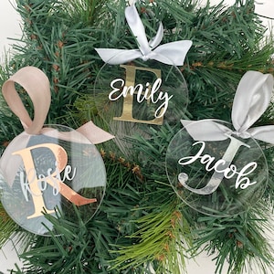 May include: Clear acrylic Christmas ornaments with names and initial letters in gold and silver. Each ornament is round and has a ribbon for hanging. The names "Emily", "Rose", and "Jacob" are displayed on the ornaments.