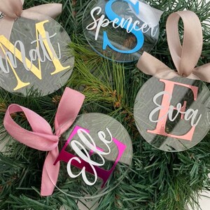 May include: Clear, round Christmas ornaments with personalized names and initials in gold, blue, pink, and rose gold. Each ornament is tied with a satin ribbon in coordinating colors. The ornaments are displayed on a bed of green pine needles.