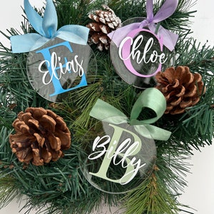 May include: Clear acrylic Christmas baubles with names "Elias", "Chloe", and "Billy" in white script. Each bauble has a coloured initial and matching ribbon. The baubles are displayed on a bed of pine needles and pine cones.