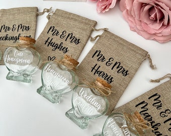 May include: Five heart-shaped glass jars with cork lids and twine bows. Each jar is filled with white sand and has a different inscription: "Our Honeymoon", "Honeymoon Sand", "Wedding Confetti", "Honeymoon Coral 2024", and "Mr & Mrs Marshall". The jars are arranged in a row on a white surface, with burlap bags behind them. The bags are personalized with the names of the couples.