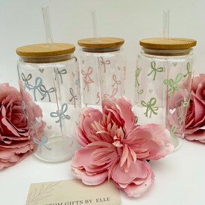 May include: Three clear glass tumblers with bamboo lids and straws, decorated with pastel bows and hearts. The tumblers are surrounded by pink flowers and a card that reads "CUSTOM GIFTS BY ELLE Created with love."