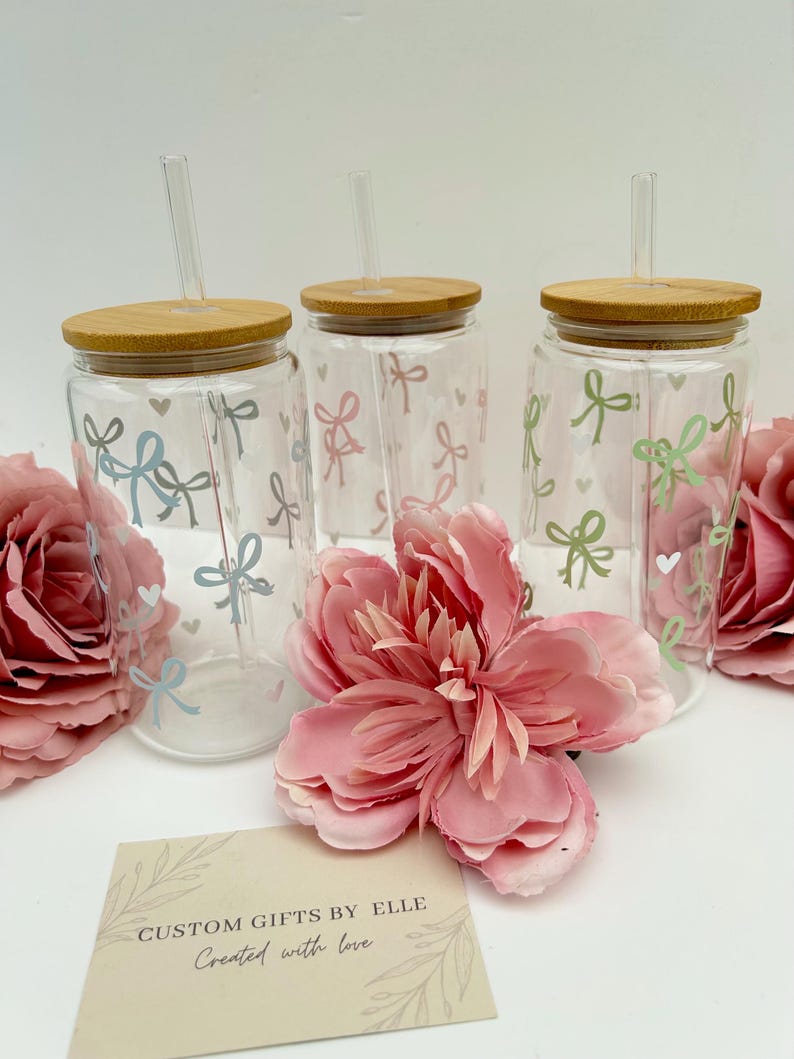 May include: Three clear glass tumblers with bamboo lids and straws, decorated with pastel bows and hearts. The tumblers are surrounded by pink flowers and a card that reads "CUSTOM GIFTS BY ELLE Created with love."