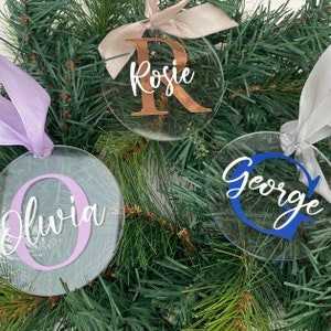 May include: Clear, round Christmas ornaments with names and initial letters. The ornaments have different colored letters and ribbons. One ornament says "Rosie" with a copper-colored "R" and a beige ribbon. Another says "Olivia" with a lavender "O" and a lavender ribbon.