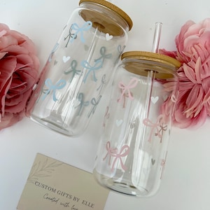 May include: Two clear glass tumblers with bamboo lids and straws, decorated with pastel bows and hearts. One tumbler has a pink bow design, the other has a blue bow design. The text "CUSTOM GIFTS BY ELLE Created with love" is on a card.