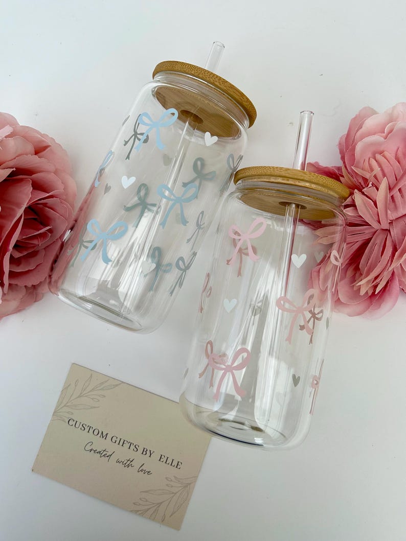 May include: Two clear glass tumblers with bamboo lids and straws, decorated with pastel bows and hearts. One tumbler has a pink bow design, the other has a blue bow design. The text "CUSTOM GIFTS BY ELLE Created with love" is on a card.