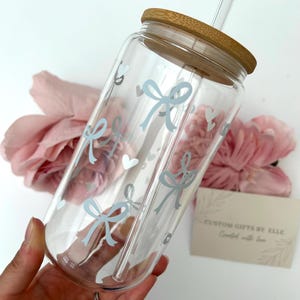 May include: A clear glass tumbler with a bamboo lid and a glass straw. The tumbler is decorated with light blue bows and hearts. The text "CUSTOM GIFTS BY ELLE Created with love" is on a small card.