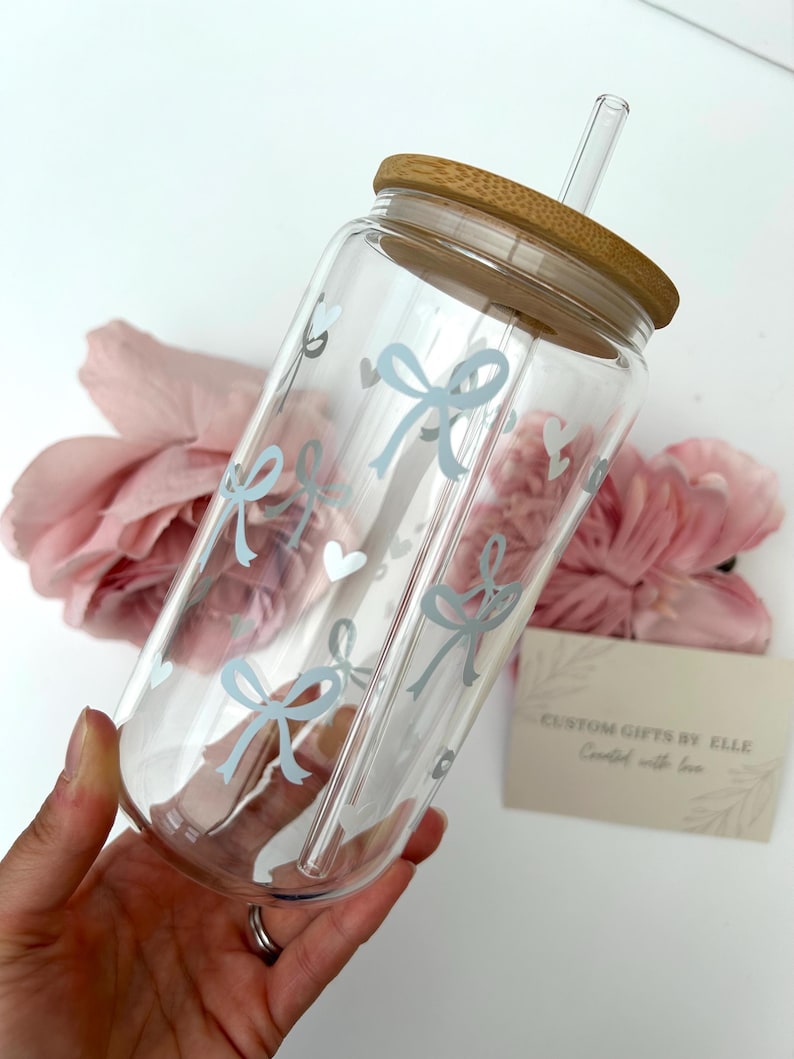 May include: A clear glass tumbler with a bamboo lid and a glass straw. The tumbler is decorated with light blue bows and hearts. The text "CUSTOM GIFTS BY ELLE Created with love" is on a small card.