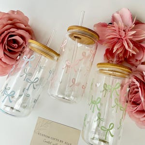 May include: Three clear glass tumblers with bamboo lids and straws, decorated with pastel bows. The bows are in shades of blue, pink, and green. The tumblers are arranged with pink flowers and a card that reads "CUSTOM GIFTS BY ELLE Created with love."