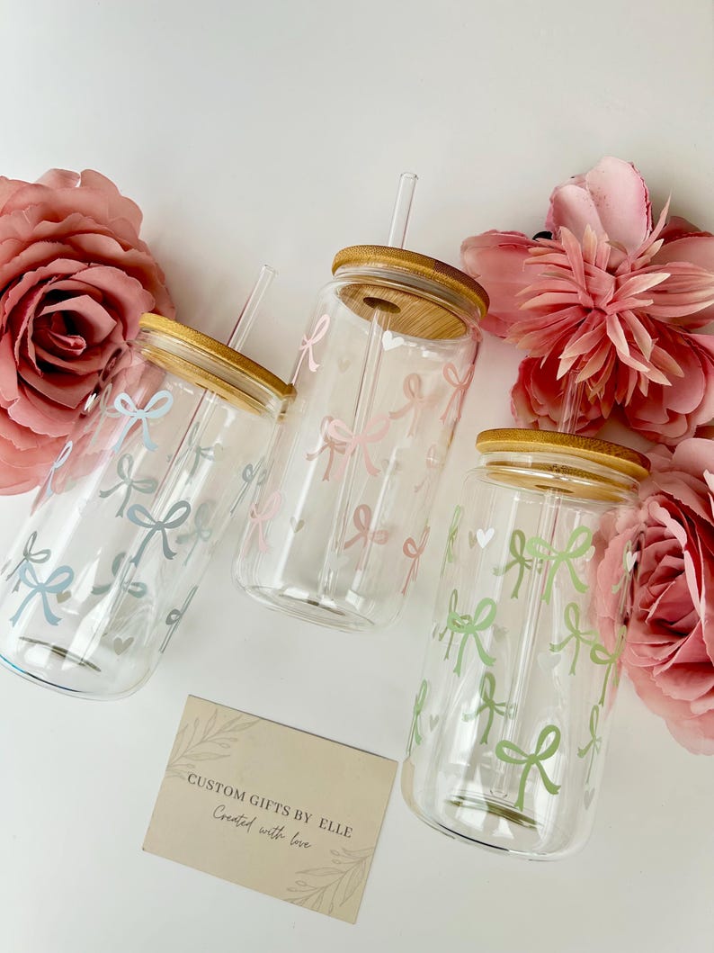May include: Three clear glass tumblers with bamboo lids and straws, decorated with pastel bows. The bows are in shades of blue, pink, and green. The tumblers are arranged with pink flowers and a card that reads "CUSTOM GIFTS BY ELLE Created with love."