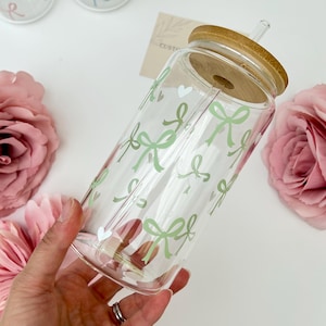 May include: Clear glass tumbler with a bamboo lid and straw, decorated with light green bows. The tumbler is held in a hand, with pink flowers in the background. The design includes small white hearts.