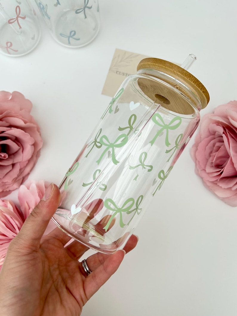 May include: Clear glass tumbler with a bamboo lid and straw, decorated with light green bows. The tumbler is held in a hand, with pink flowers in the background. The design includes small white hearts.