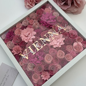 May include: A white frame filled with pink and purple paper flowers. The name "Vienna" is written in gold letters on the flowers.