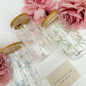 May include: Clear glass tumblers with bamboo lids and straws, decorated with pastel bows. The tumblers are arranged with pink flowers and a card that reads "CUSTOM GIFTS BY ELLE Created with love."