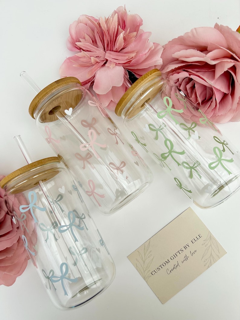 May include: Clear glass tumblers with bamboo lids and straws, decorated with pastel bows. The tumblers are arranged with pink flowers and a card that reads "CUSTOM GIFTS BY ELLE Created with love."