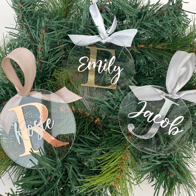 May include: Clear acrylic Christmas ornaments with names and initial letters in gold and silver. Each ornament is round and has a ribbon for hanging. The names "Emily", "Rose", and "Jacob" are displayed on the ornaments.
