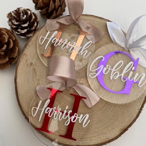 May include: Clear acrylic personalized name ornaments with white script names and colored initial letters. Each ornament has a satin ribbon for hanging. The names are "Hannah", "Goblin", and "Harrison". The ornaments are displayed on a wooden slice with pinecones.