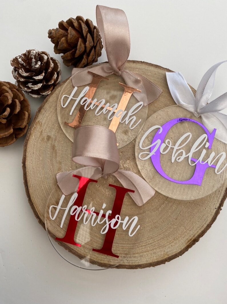 May include: Clear acrylic personalized name ornaments with white script names and colored initial letters. Each ornament has a satin ribbon for hanging. The names are "Hannah", "Goblin", and "Harrison". The ornaments are displayed on a wooden slice with pinecones.