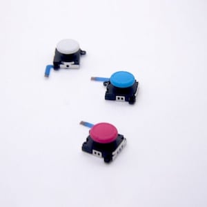 Könnte beinhalten: Drei Ersatz-Joystick-Knöpfe für einen Videospiel-Controller. Die Knöpfe sind weiß, blau und rosa.