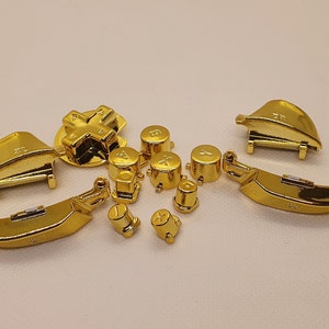 Könnte beinhalten: Ein Satz goldfarbener Ersatztasten für einen Videospielcontroller. Das Set enthält ein Richtungskreuz, vier Gesichtstasten mit den Beschriftungen A, B, X und Y sowie zwei Schultertasten.