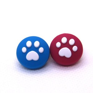 Könnte beinhalten: Zwei Joystick-Kappen in Form von Pfotenabdrücken, eine blaue und eine rote, mit weißen Pfotenabdrücken.