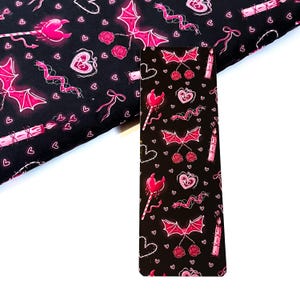 May include: Black fabric with a repeating pattern of pink bats, hearts, roses, and other gothic-themed elements. A bookmark with the same pattern is also visible. The design is suitable for crafting projects.