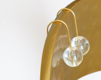 May include: A pair of gold hoop earrings with clear glass beads. The earrings are hanging on a gold surface.