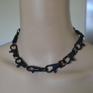 May include: A black necklace made from multiple clasp connectors. The necklace is displayed on a mannequin. The connectors are evenly spaced, creating a bold, industrial look. The necklace is a dark, matte black.