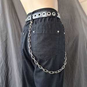 May include: Black denim pants with a silver chain attached to a black leather belt with silver grommets. The chain hangs down from the belt loop to the back pocket of the pants.
