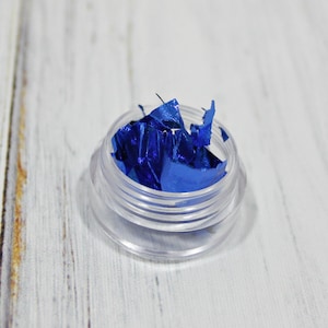 May include: A small, clear plastic container filled with vibrant blue metallic flakes. The container sits on a white wooden surface, creating a contrast between the bright blue and the neutral background. The flakes appear to be irregularly shaped.